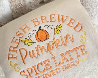 May include: Cream-colored rectangular pillow with embroidered text: "FRESH BREWED Pumpkin SPICE LATTE SERVED DAILY." The design includes an orange pumpkin and yellow leaves. The pillow is on a white, textured surface.