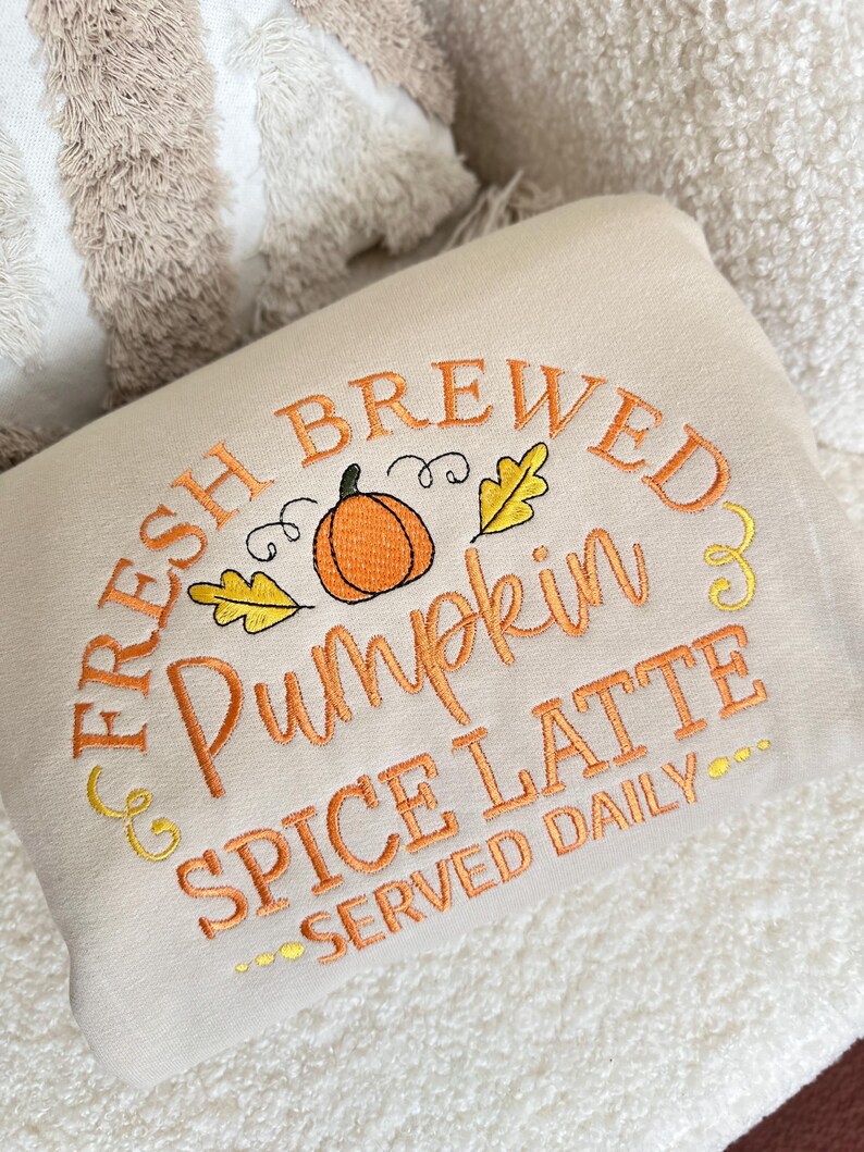 Pode incluir: Almofada retangular creme com texto bordado: "FRESH BREWED Pumpkin SPICE LATTE SERVED DAILY." O design inclui uma ab&oacute;bora laranja e folhas amarelas. A almofada est&aacute; sobre uma superf&iacute;cie branca texturizada.