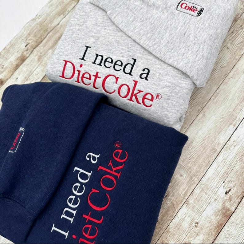 Puede incluir: Dos sudaderas, una gris y otra azul marino, bordadas con "I need a Diet Coke" en letras rojas y blancas. La sudadera gris tiene un peque&ntilde;o logotipo de "Coke". La sudadera azul marino est&aacute; doblada, mostrando el texto.