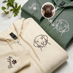 Puede incluir: Una sudadera con cremallera color crema con una ilustración de líneas marrones de una cara de perro y el nombre Buddy con una huella en la manga. Al lado, una sudadera verde salvia muestra un dibujo lineal blanco de un perro y una foto.