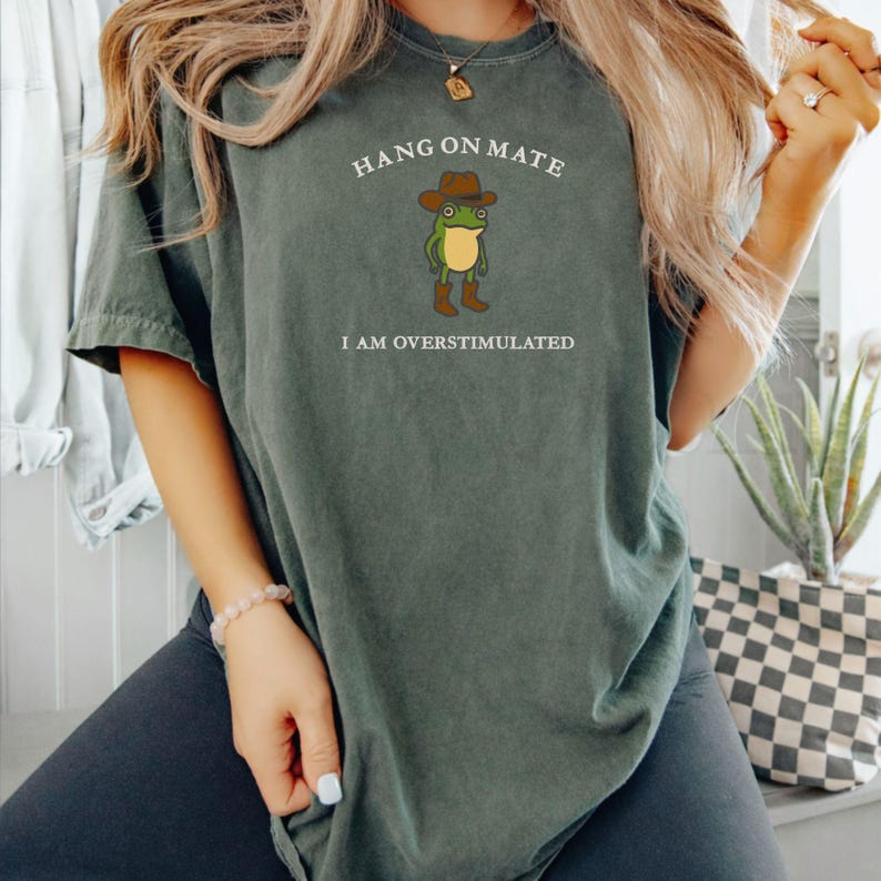 May include: Dark green t-shirt with the text "HANG ON MATE" above a cartoon frog wearing a cowboy hat and boots. The text "I AM OVERSTIMULATED" is below the frog. The shirt is a relaxed fit.