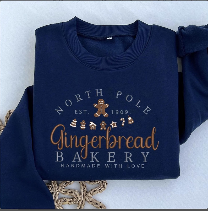 May include: Navy blue sweatshirt with embroidered text and gingerbread-themed graphics. The design includes "NORTH POLE EST. 1909" above "Gingerbread Bakery" and "HANDMADE WITH LOVE". Features a gingerbread man and other festive icons.