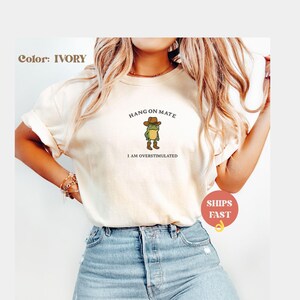 May include: Ivory-colored t-shirt featuring a cartoon frog wearing a cowboy hat and boots. The text above the frog reads "HANG ON MATE," and below it, "I AM OVERSTIMULATED." The shirt is a casual, comfortable style.