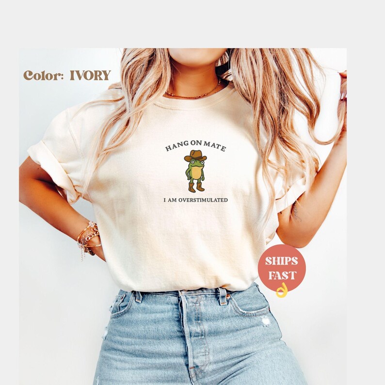 May include: Ivory-colored t-shirt featuring a cartoon frog wearing a cowboy hat and boots. The text above the frog reads "HANG ON MATE," and below it, "I AM OVERSTIMULATED." The shirt is a casual, comfortable style.