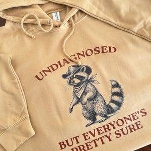 May include: A tan hoodie featuring a raccoon wearing a cowboy hat and bandana. The text "UNDIAGNOSED BUT EVERYONE'S PRETTY SURE" is printed in red on the hoodie.