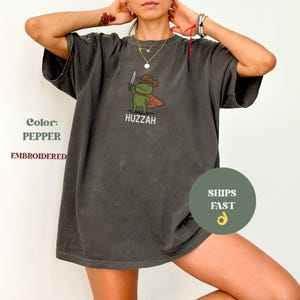 May include: Pepper-colored, embroidered t-shirt featuring a cartoon frog in a cowboy hat and cape, holding a knife. The word "Huzzah" is below the frog. The shirt has a relaxed fit.