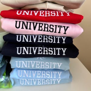 May include: A stack of sweatshirts in various colors, each with the word "UNIVERSITY" embroidered in white on the front.