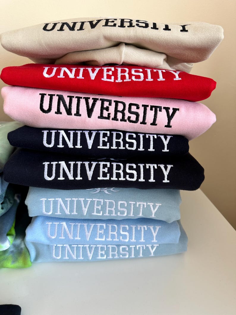 May include: A stack of sweatshirts in various colors, each with the word "UNIVERSITY" embroidered in white on the front.