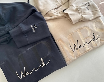 May include: Two hooded sweatshirts, one charcoal gray and one beige, each featuring large embroidered lettering that reads "MR" or "MRS" with the cursive name "Ward" overlaid. The sleeve of each hoodie is embroidered with "EST 19.2025".