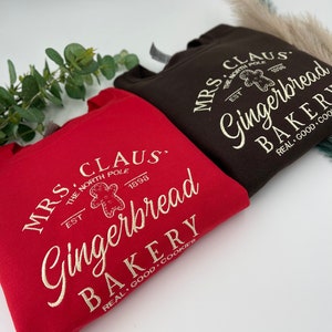 May include: Two sweatshirts, one red and one brown, with the text "MRS. CLAUS Gingerbread BAKERY" in a vintage style. The design includes a gingerbread man graphic and the words "REAL GOOD COOKIES".