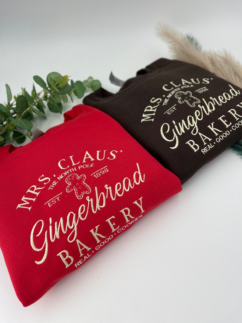 May include: Two sweatshirts, one red and one brown, with the text "MRS. CLAUS Gingerbread BAKERY" in a vintage style. The design includes a gingerbread man graphic and the words "REAL GOOD COOKIES".
