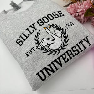May include: Gray sweatshirt with black embroidered text that reads "Silly Goose University Est. 1910" and a white swan with a gold crown.