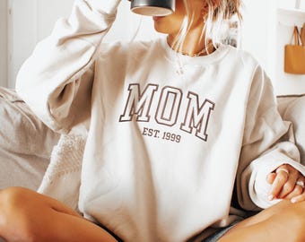 May include: A cream colored sweatshirt with the text "MOM EST. 1999" in black.