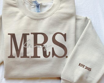 May include: A beige sweatshirt with the word "MRS" in brown letters and the word "EST 2025" in brown letters on the sleeve.