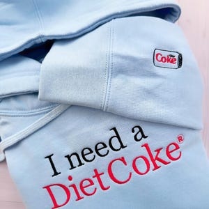 Puede incluir: Sudadera con capucha azul claro con la frase bordada "I need a Diet Coke" en negro y rojo. La capucha presenta una peque&ntilde;a lata de Diet Coke bordada. La sudadera tiene un cord&oacute;n y un aspecto suave y c&oacute;modo.