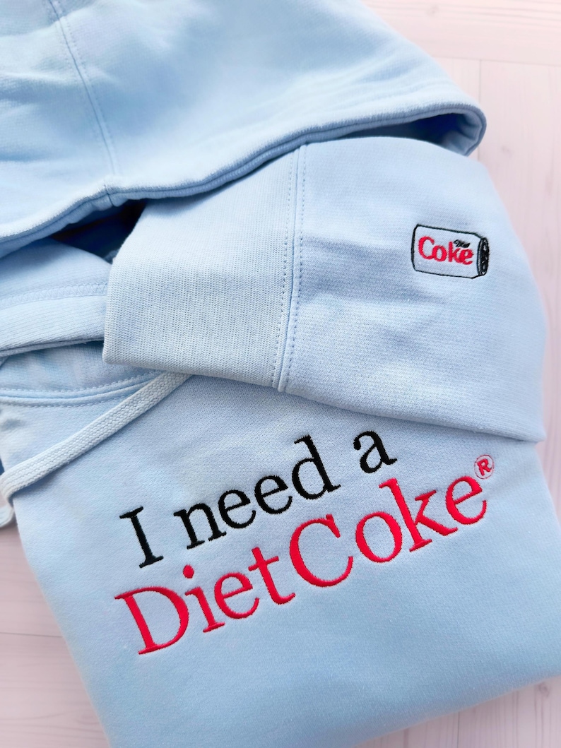 Puede incluir: Sudadera con capucha azul claro con la frase bordada "I need a Diet Coke" en negro y rojo. La capucha presenta una peque&ntilde;a lata de Diet Coke bordada. La sudadera tiene un cord&oacute;n y un aspecto suave y c&oacute;modo.
