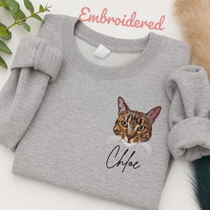 May include: A light gray sweatshirt with an embroidered cat face and the name "Chloe" in cursive. The cat has brown and tan fur with pink ears and yellow eyes. The word "Embroidered" is written in pink script at the top.