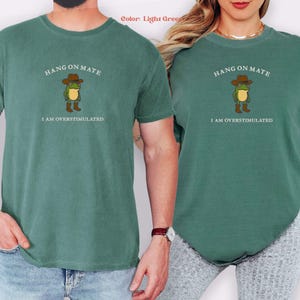 May include: Light green t-shirts with the text "HANG ON MATE" above a cartoon frog wearing a cowboy hat and boots. Below the frog is the text "I AM OVERSTIMULATED". The shirts are a solid color.