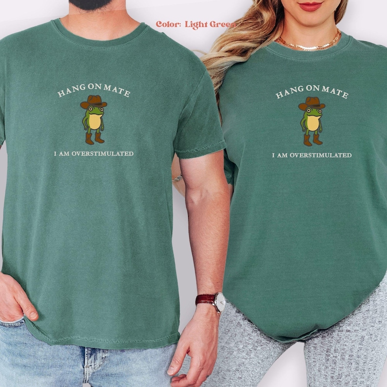May include: Light green t-shirts with the text "HANG ON MATE" above a cartoon frog wearing a cowboy hat and boots. Below the frog is the text "I AM OVERSTIMULATED". The shirts are a solid color.