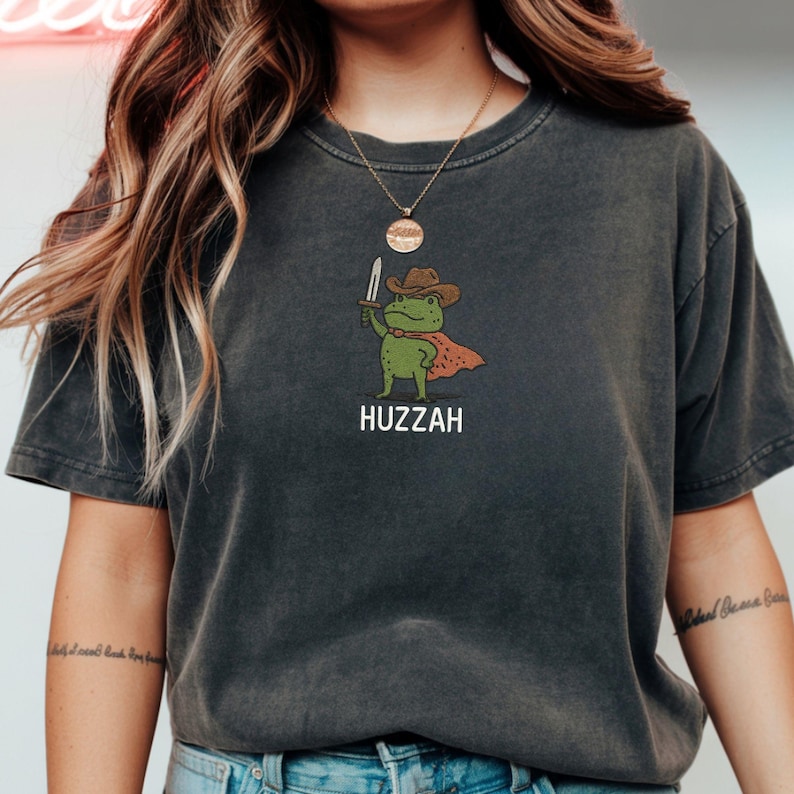 May include: Dark gray t-shirt featuring a cartoon frog wearing a cowboy hat and cape, holding a sword. The word "Huzzah" is printed below the frog illustration. The shirt has a vintage, worn look.