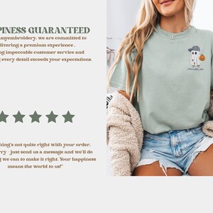 May include: A light green t-shirt featuring an embroidered ghost wearing a baseball cap and holding a pumpkin. The shirt has the text "HAPPINESS GUARANTEED" and a customer satisfaction message.