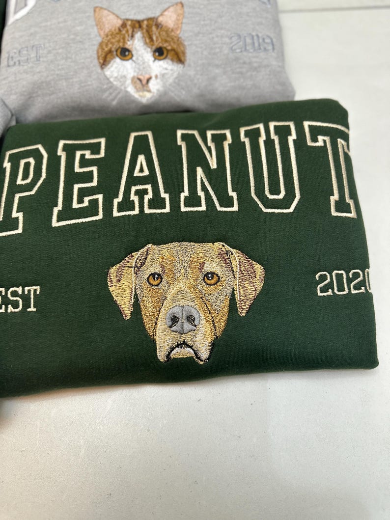 May include: Green sweatshirt with the name "PEANUT" embroidered in white block letters above a detailed dog face. The year "2020" is embroidered on the right side. A gray sweatshirt with a cat face and the year "2019" is visible in the background.