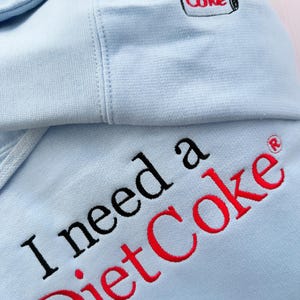 Puede incluir: Sudadera con capucha azul claro con la frase bordada "I need a Diet Coke" en negro y rojo. La capucha presenta un peque&ntilde;o logotipo de "Coke" bordado. La sudadera est&aacute; hecha de un tejido suave.