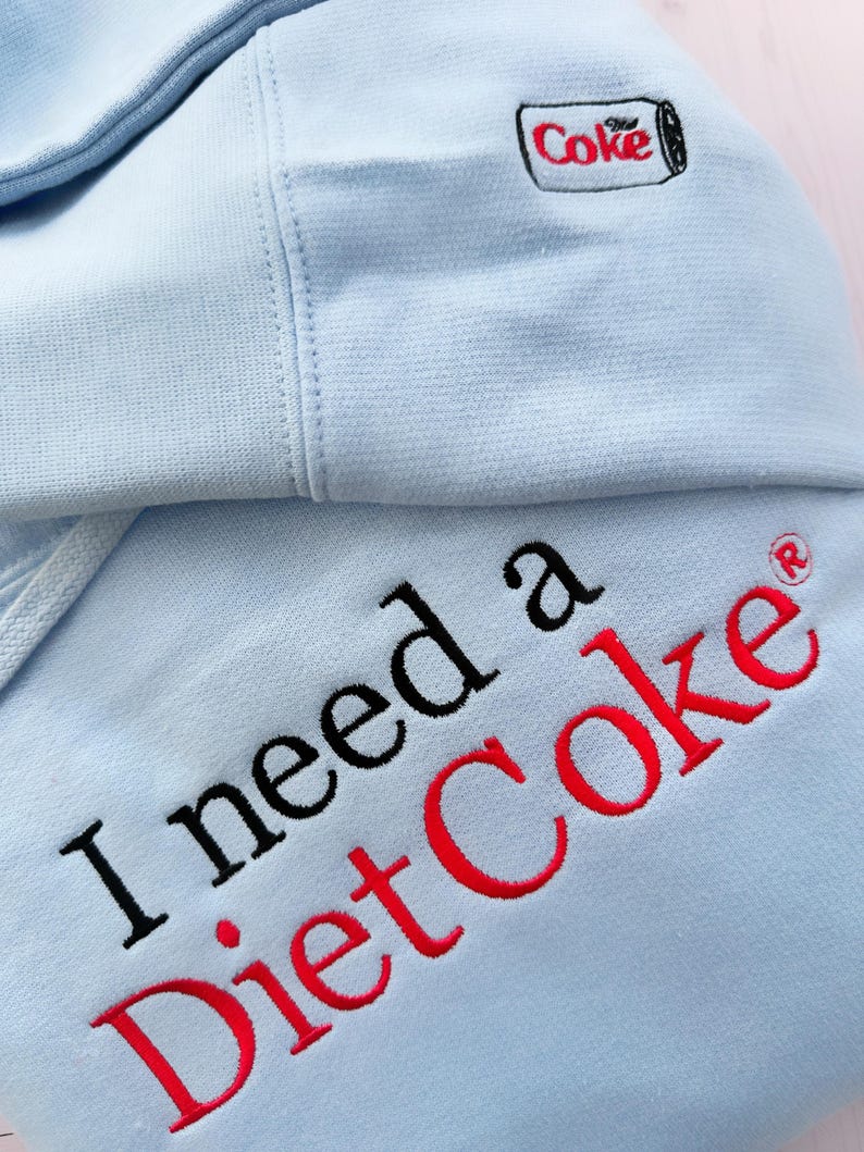 Puede incluir: Sudadera con capucha azul claro con la frase bordada "I need a Diet Coke" en negro y rojo. La capucha presenta un peque&ntilde;o logotipo de "Coke" bordado. La sudadera est&aacute; hecha de un tejido suave.