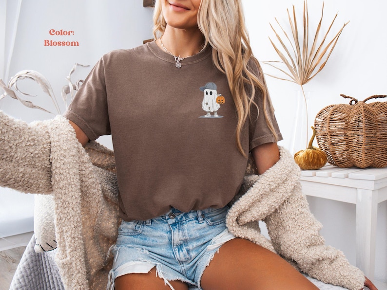 May include: A brown t-shirt with a small embroidered design of a ghost wearing a baseball cap and holding a pumpkin. The shirt is a solid color and has short sleeves. The text "Color: Blossom" is in the upper left corner.