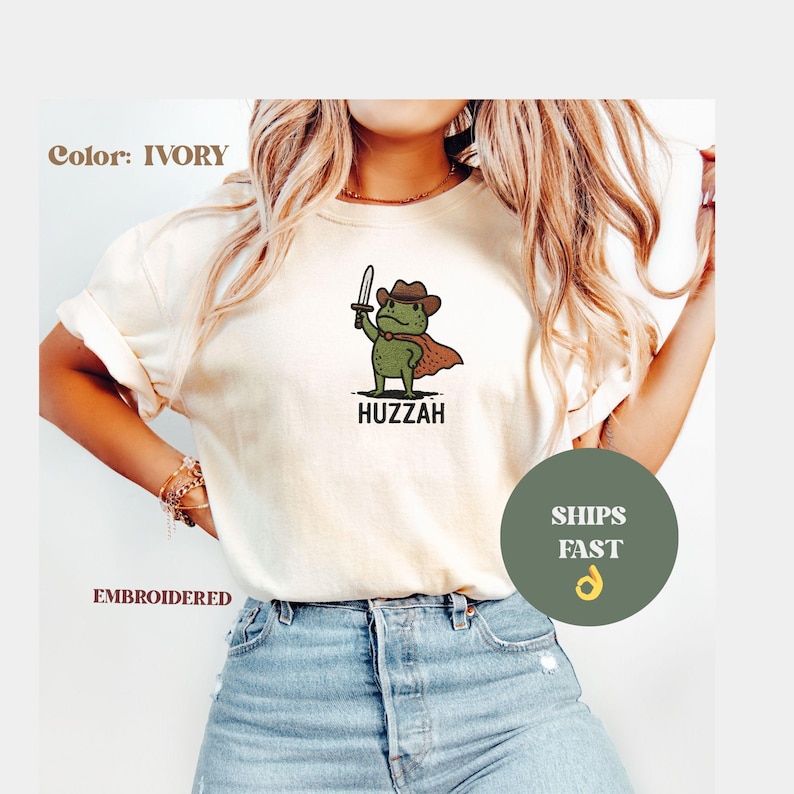May include: Ivory-colored t-shirt with an embroidered design. The graphic features a green frog wearing a cowboy hat and cape, holding a sword, with the word "Huzzah" below. The shirt is a casual wear item.