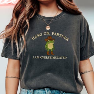 Puede incluir: Camiseta gris oscuro con el texto "HANG ON, PARTNER" sobre una rana de dibujos animados con sombrero y botas de vaquero. El texto "I AM OVERSTIMULATED" está debajo. La camiseta es de estilo casual.