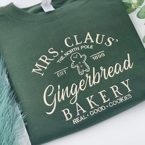May include: Dark green sweatshirt with the text "MRS. CLAUS, THE NORTH POLE, EST. 1898 Gingerbread BAKERY REAL GOOD COOKIES" in a cream-colored font. The design includes a gingerbread man graphic.
