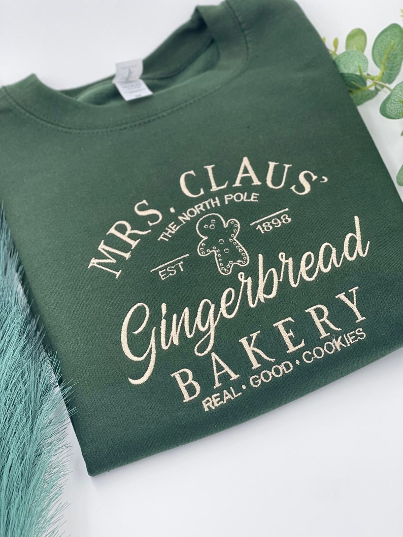 May include: Dark green sweatshirt with the text "MRS. CLAUS, THE NORTH POLE, EST. 1898 Gingerbread BAKERY REAL GOOD COOKIES" in a cream-colored font. The design includes a gingerbread man graphic.