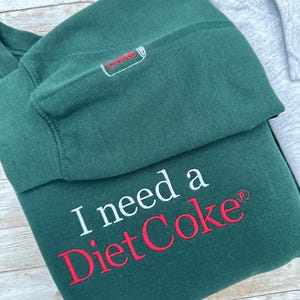 Puede incluir: Una sudadera verde oscuro con la frase "I need a Diet Coke" bordada en blanco y rojo. La manga presenta una peque&ntilde;a lata de Diet Coke bordada. Una sudadera gris es parcialmente visible.