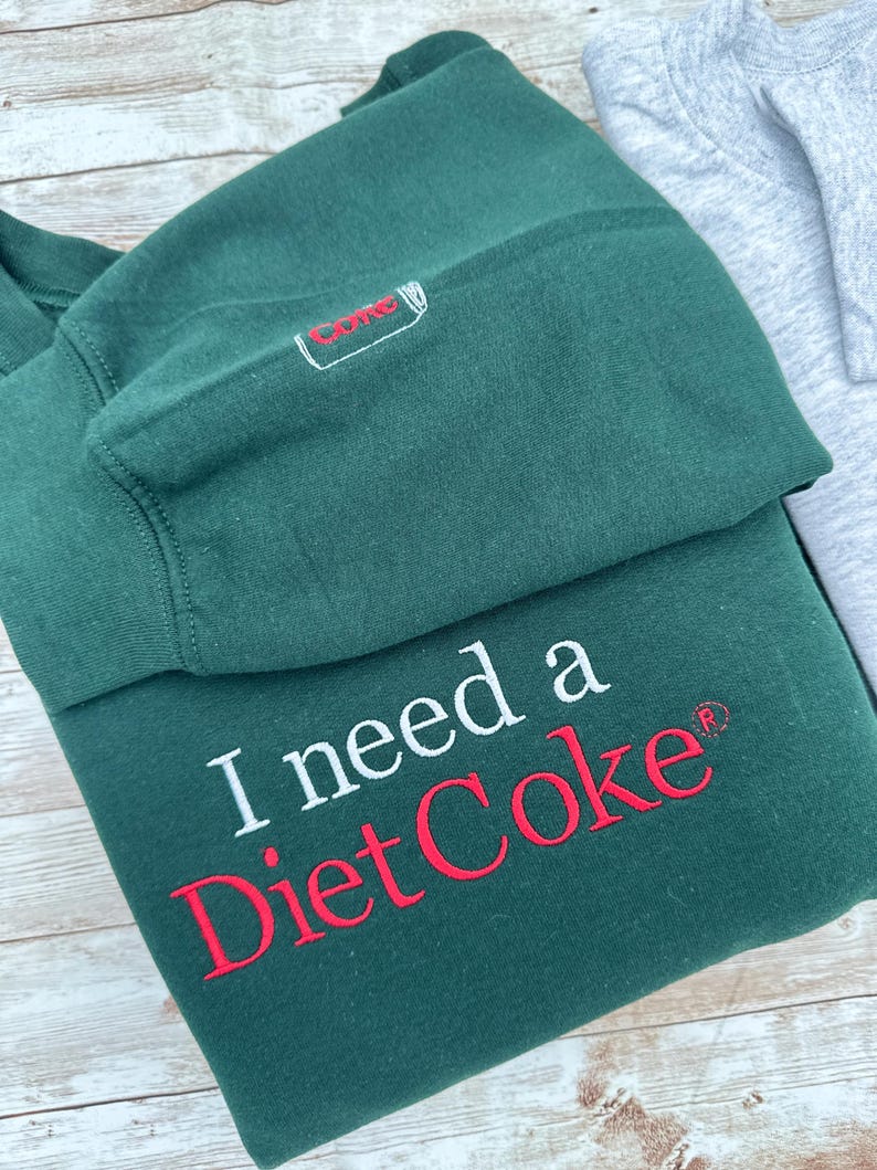 Puede incluir: Una sudadera verde oscuro con la frase "I need a Diet Coke" bordada en blanco y rojo. La manga presenta una peque&ntilde;a lata de Diet Coke bordada. Una sudadera gris es parcialmente visible.