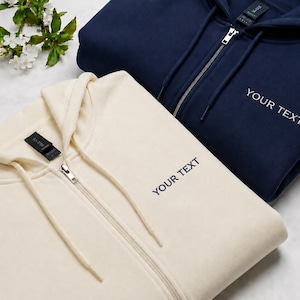 Può includere: Una felpa con cappuccio e zip color crema e una felpa con cappuccio e zip blu navy, entrambe dotate di cappuccio con coulisse abbinate e la scritta "YOUR TEXT" ricamata sul petto a sinistra. Le felpe hanno una chiusura a zip in metallo.