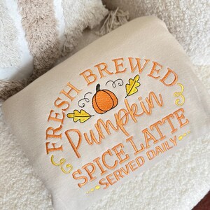 Pode incluir: Uma almofada bege bordada com o texto "FRESH BREWED Pumpkin SPICE LATTE SERVED DAILY" em fio laranja. O desenho inclui uma ab&oacute;bora e folhas. A almofada est&aacute; sobre uma superf&iacute;cie branca e texturizada.