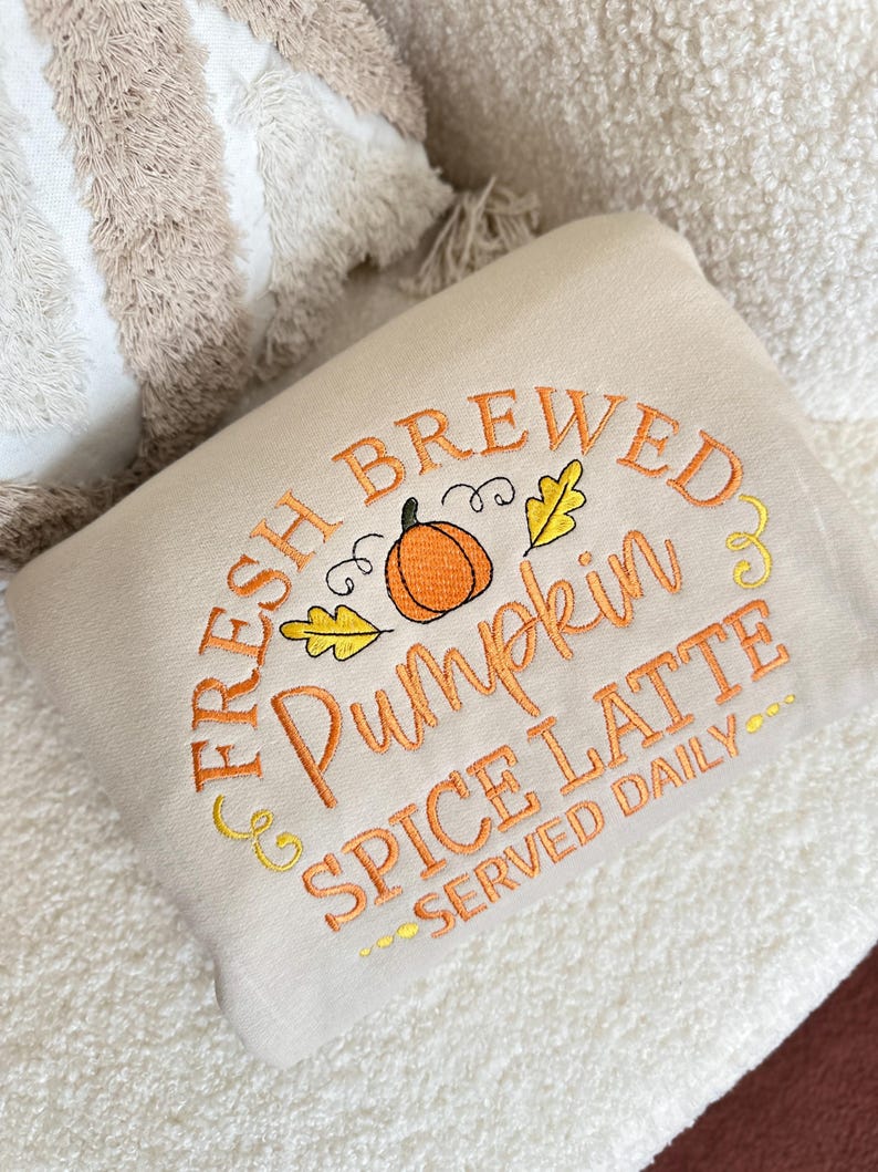 Pode incluir: Uma almofada bege bordada com o texto "FRESH BREWED Pumpkin SPICE LATTE SERVED DAILY" em fio laranja. O desenho inclui uma ab&oacute;bora e folhas. A almofada est&aacute; sobre uma superf&iacute;cie branca e texturizada.