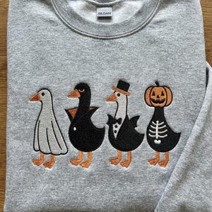 May include: A grey sweatshirt with an embroidered design of four cartoon ducks in Halloween costumes. The ducks are dressed as a ghost, vampire, in a tuxedo, and a jack-o'-lantern. The sweatshirt is made of a soft, comfortable material.