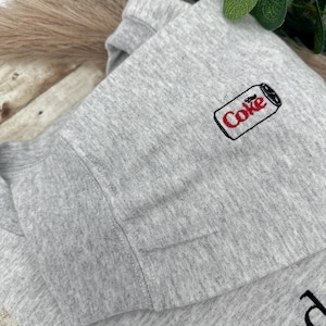 Puede incluir: Sudadera gris claro con un peque&ntilde;o logotipo bordado de una lata de Coca-Cola en la parte superior izquierda del pecho. La palabra "Coke" es roja con un contorno negro. La sudadera est&aacute; doblada.