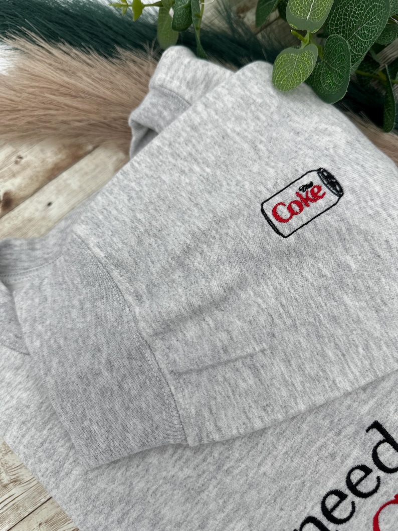 Puede incluir: Sudadera gris claro con un peque&ntilde;o logotipo bordado de una lata de Coca-Cola en la parte superior izquierda del pecho. La palabra "Coke" es roja con un contorno negro. La sudadera est&aacute; doblada.