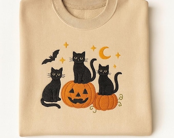 Puede incluir: Sudadera beige con un diseño bordado de Halloween. El diseño presenta tres gatos negros sentados sobre calabazas naranjas, con una luna creciente, estrellas y un murciélago. La sudadera tiene cuello redondo y mangas largas.