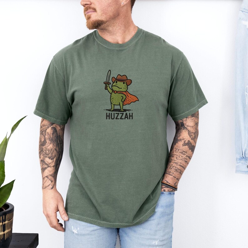 May include: Olive green t-shirt featuring a cartoon frog wearing a cowboy hat and cape, holding a sword. The word "Huzzah" is printed below the frog. The shirt is a solid color and has short sleeves.