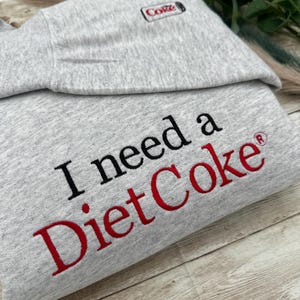 Puede incluir: Sudadera gris claro con el texto "I need a Diet Coke" bordado en negro y rojo. La esquina superior izquierda presenta un peque&ntilde;o logotipo de Diet Coke bordado. La sudadera est&aacute; doblada sobre una superficie de madera.