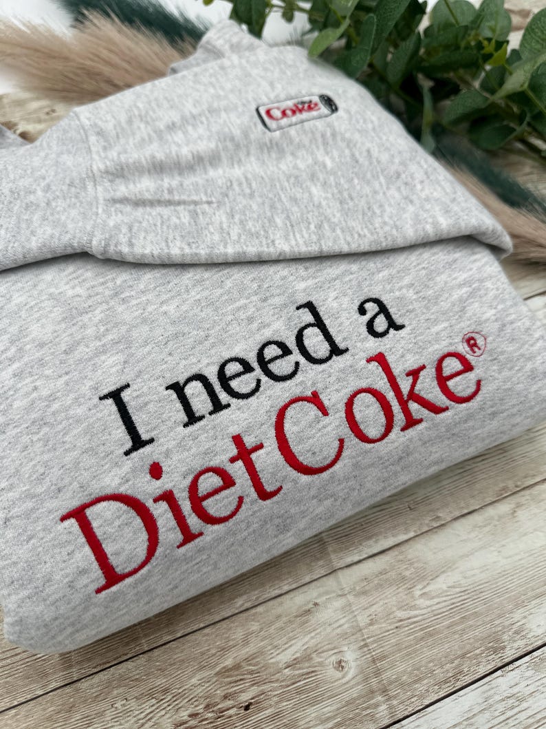 Puede incluir: Sudadera gris claro con el texto "I need a Diet Coke" bordado en negro y rojo. La esquina superior izquierda presenta un peque&ntilde;o logotipo de Diet Coke bordado. La sudadera est&aacute; doblada sobre una superficie de madera.