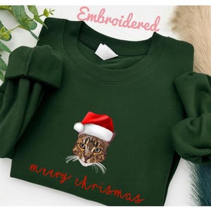 Puede incluir: Una sudadera verde oscuro con un gato bordado con un gorro de Papá Noel. Las palabras "merry chrismas" están escritas en rojo debajo del gato. La palabra "Embroidered" está escrita en rosa encima de la sudadera.
