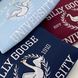 May include: Three sweatshirts with different colored backgrounds and white embroidered text. The text reads "Silly Goose University" and includes a goose graphic. The sweatshirts are blue, maroon, and navy blue.