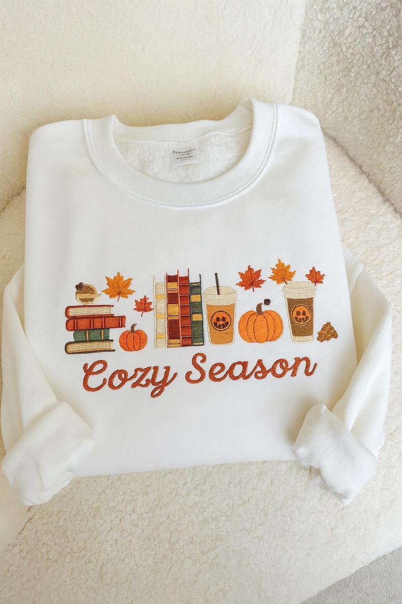 May include: White sweatshirt with an embroidered design featuring books, pumpkins, fall leaves, and coffee cups. The phrase "Cozy Season" is embroidered in orange script below the design. The sweatshirt has a crew neck and long sleeves.