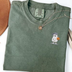 May include: A moss green t-shirt with a small embroidered ghost wearing a hat and holding a pumpkin. A gold necklace and a brown hat are also visible. The shirt has a Comfort Colors tag.