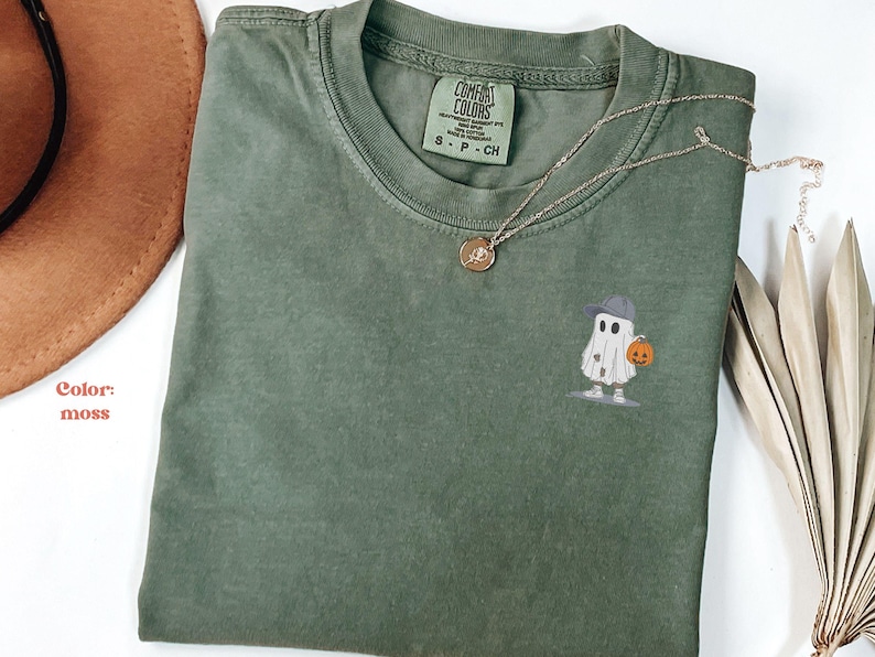 May include: A moss green t-shirt with a small embroidered ghost wearing a hat and holding a pumpkin. A gold necklace and a brown hat are also visible. The shirt has a Comfort Colors tag.